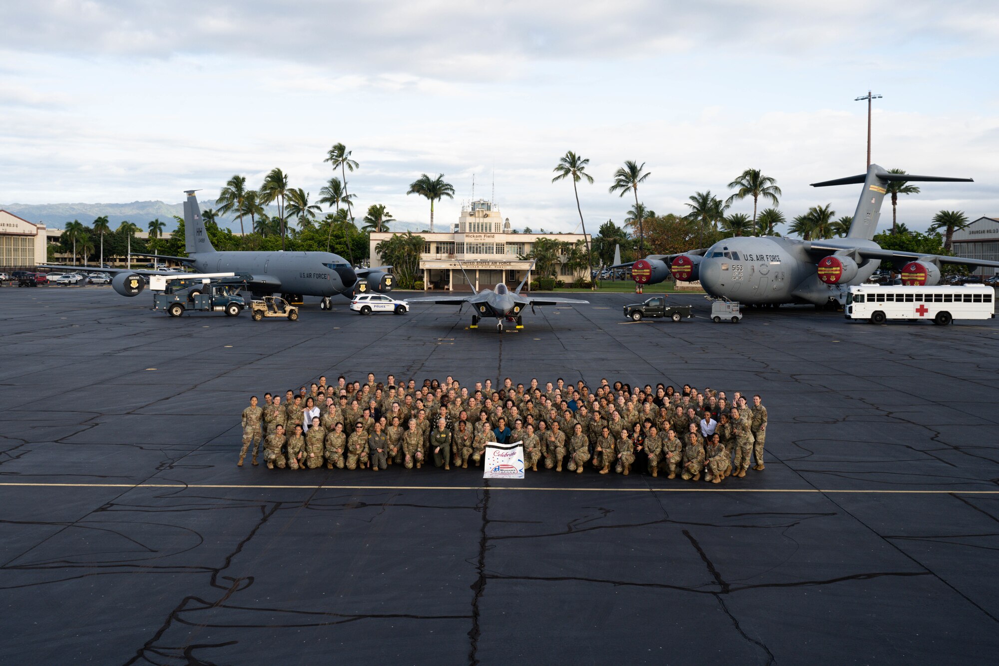 Women assigned to Joint Base Pearl Harbor Hickam stand for a group photo with static aircraft in the background