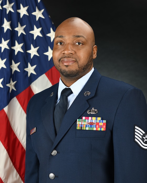 Official Photo of TSgt David Dormeus, arranger and composer with the USAF Band of the Pacific-Asia, Yokota Air Base, Japan.  TSgt Dormeus is wearing blue service dress in front of an American flag background.