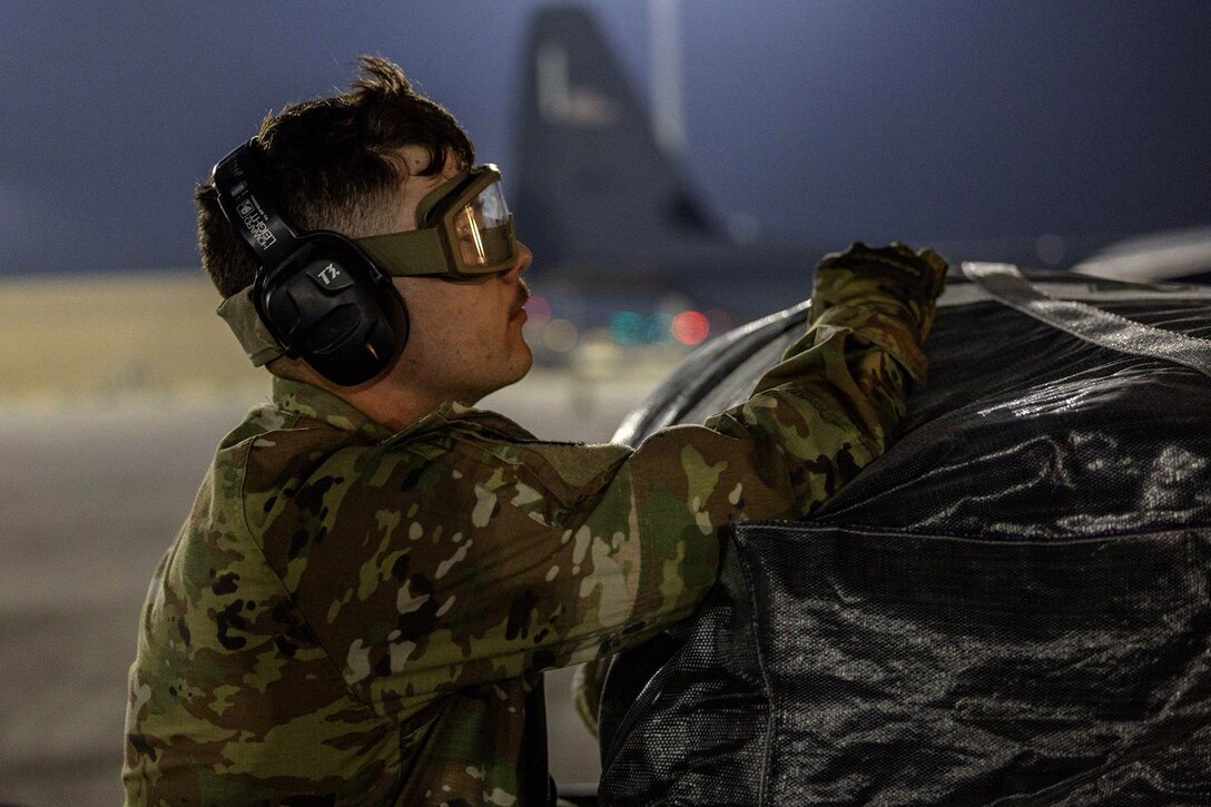 An airman adjusts a strap on a large package sitting on a flight line, with a darkened sky as backdrop.