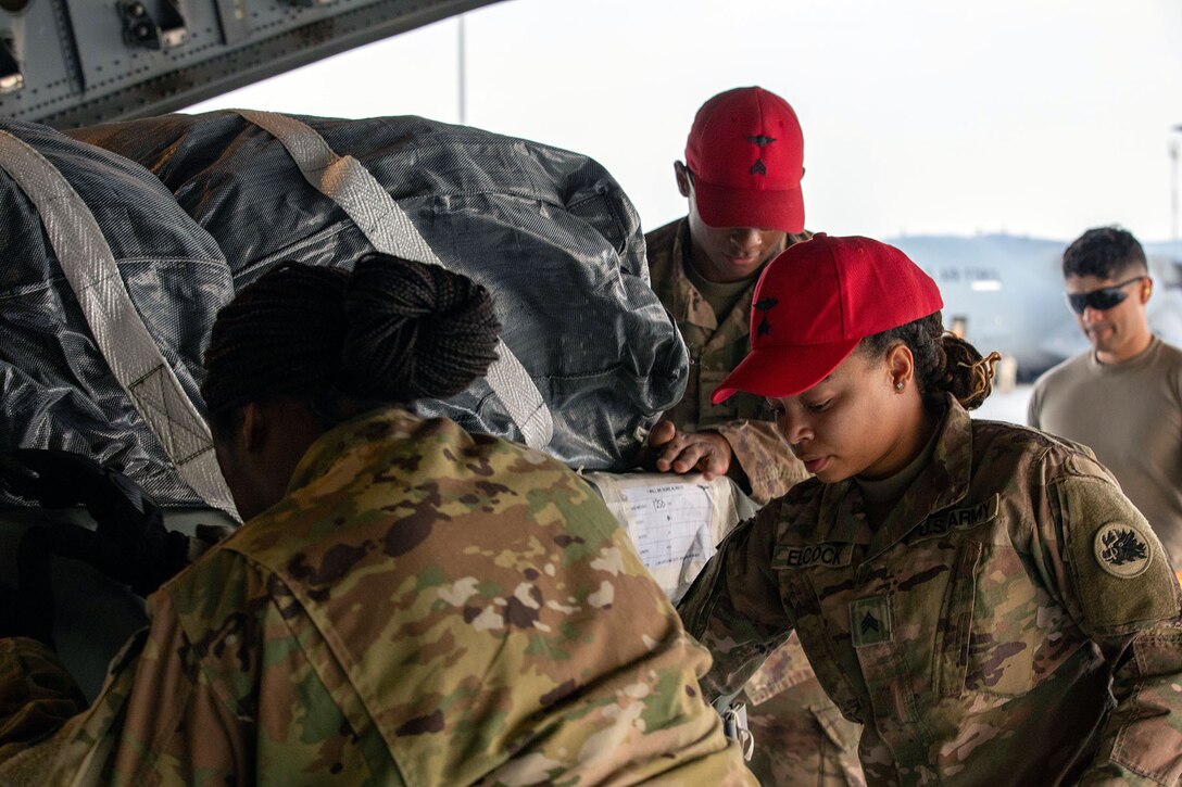 Soldiers, including two wearing red baseball hats, handle cargo on a flight line.
