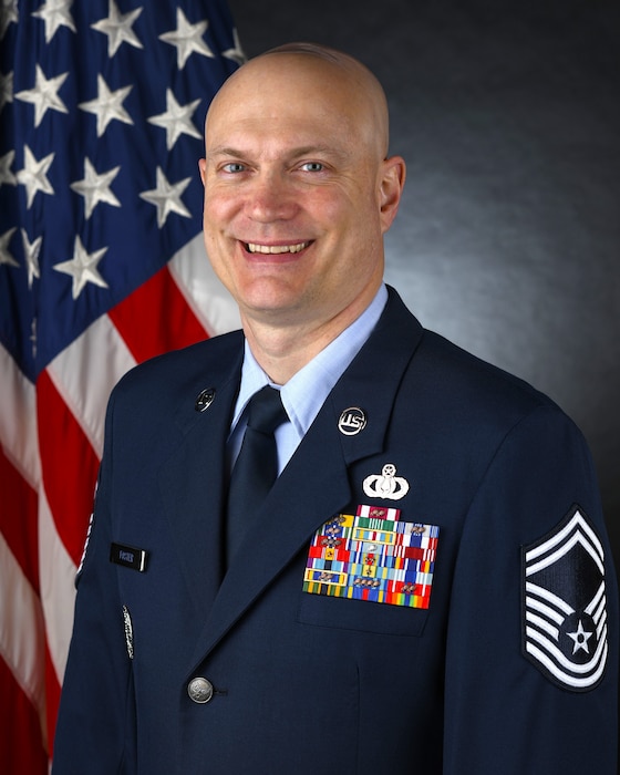 Official Photo of SMSgt Jason Foster, Senior Enlisted Leader for the USAF Band of the Pacific.  SMSgt Foster is wearing blue service dress in front of the American flag.