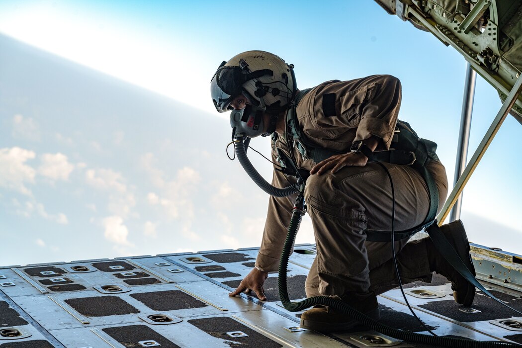 A Marine wearing a flight suit and oxygen mask kneels on the open back ramp of a cargo aircraft.