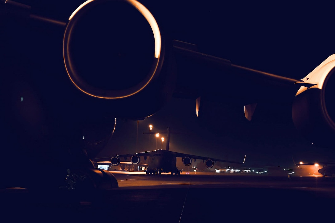 The engines of a large aircraft are seen as it sits on the tarmac of a lit airfield at night. Another aircraft is behind it.