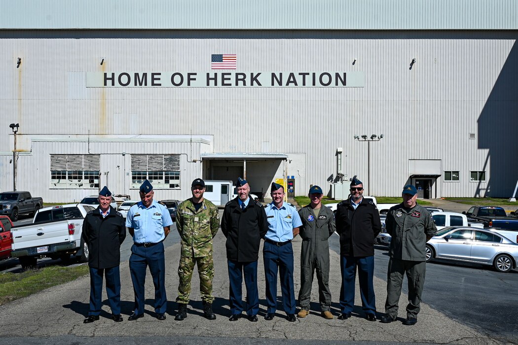 Uniformed personnel stand together in front of an aircraft hanger