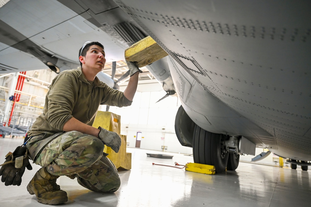 A kneeling airman wedges a piece of wood into a small door under the wing of an aircraft in a garage.