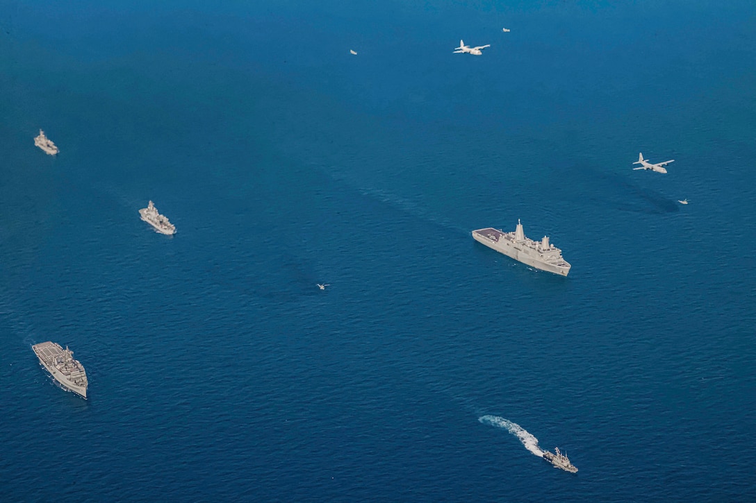 Nine ships sail in formation in a body of water as two aircraft fly overhead as seen from above.