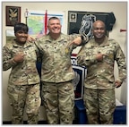 An Army Lt. Col., Command Sgt. Maj. and Sgt. Maj. pose pointing to their recruiting gold badge on their uniform.