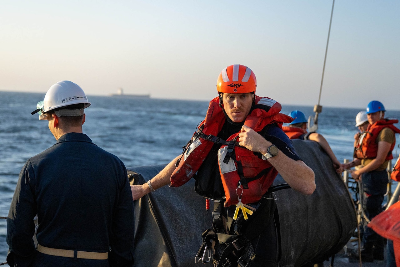A person in coveralls wearing a life jacket and helmet is facing the camera while checking the life jacket. A person in dark coveralls with their back to the camera is to the left. The ocean is in the background with a large cargo ship in the distance.