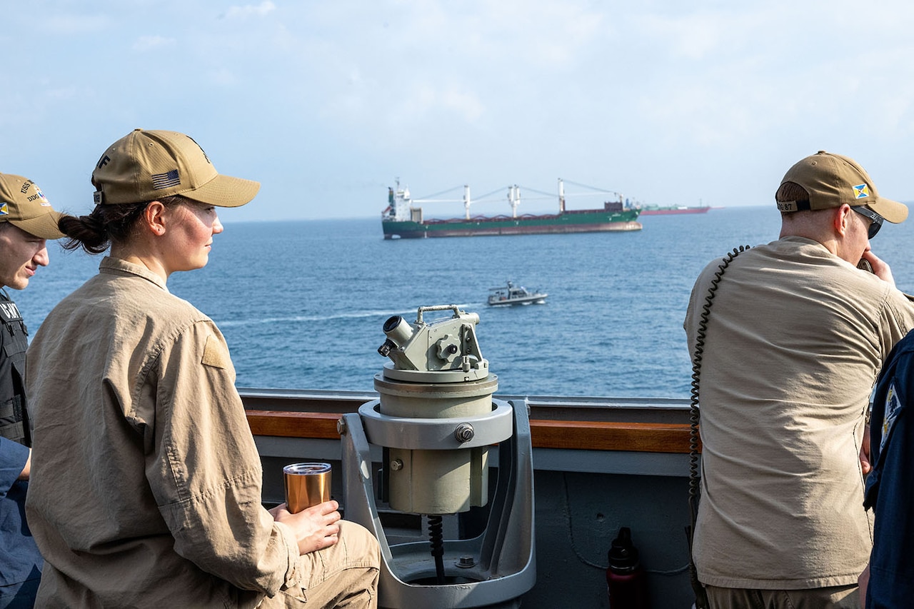 Three people wearing coveralls stand outside on the deck of a ship and look at a cargo vessel in the distance.