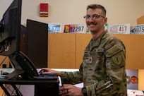 Spc. Ethan Moorhouse, human resources specialist with 54th Troop Command, New Hampshire Army National Guard, poses for a photo at his workstation at the Edward Cross Training Complex in Pembroke, N.H. Moorhouse, known by his section as "Powerhouse" was responsible for the successful processing of 1,418 Customer Relationship Management cases across the National Guard in 2023.