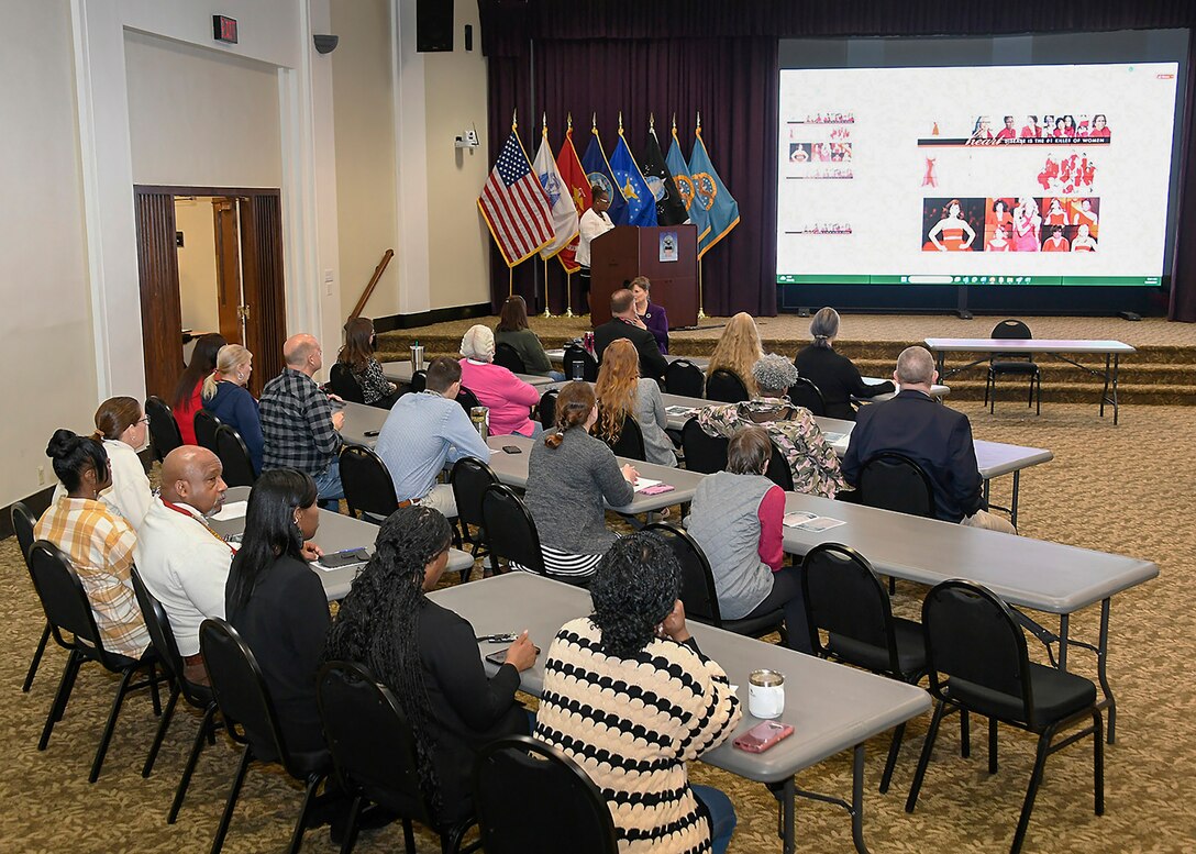 A diverse group of adults sitting in chairs at tables are seen from the back. they are looking at a person at a podium and a large TV screen with a information slide on it.