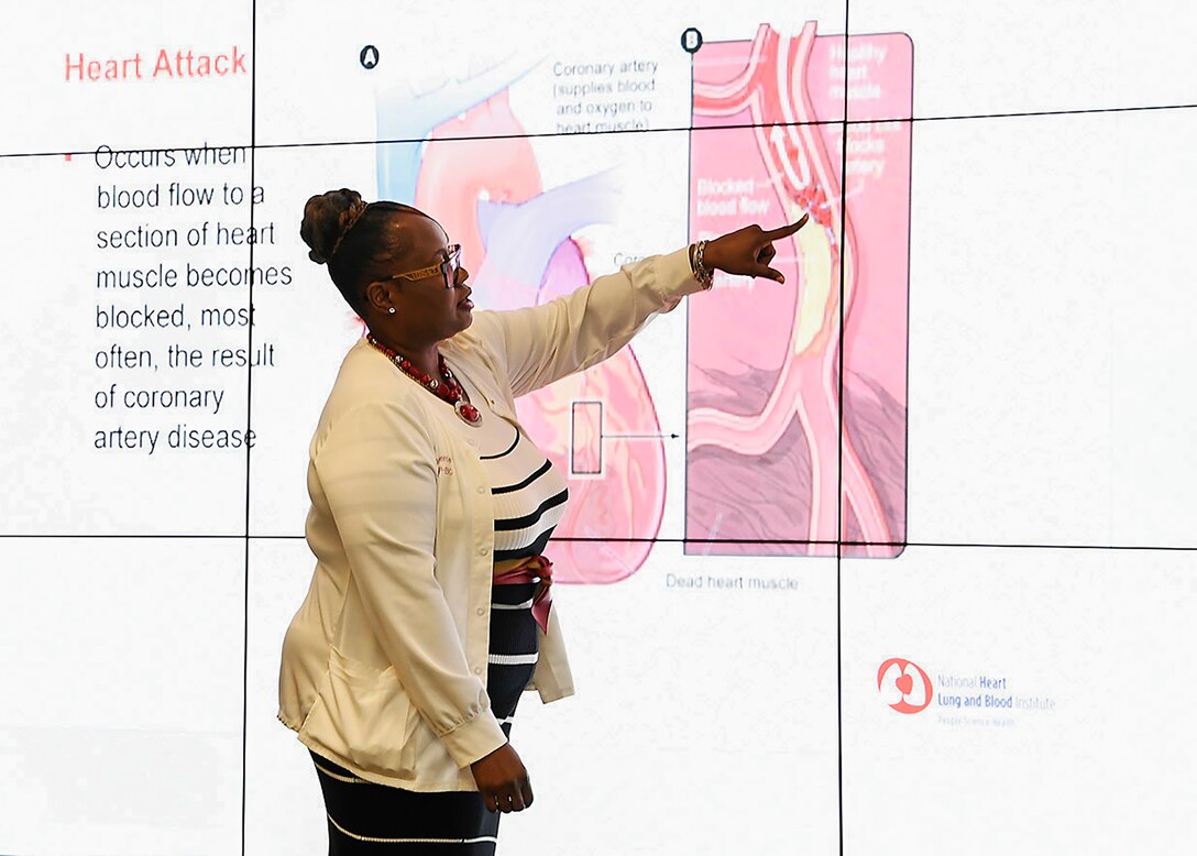 A woman stands in front of large image of a heart. she is pointing at an area that represents heart disease. Some of the text on the slide is readable and says "Heart Attack".