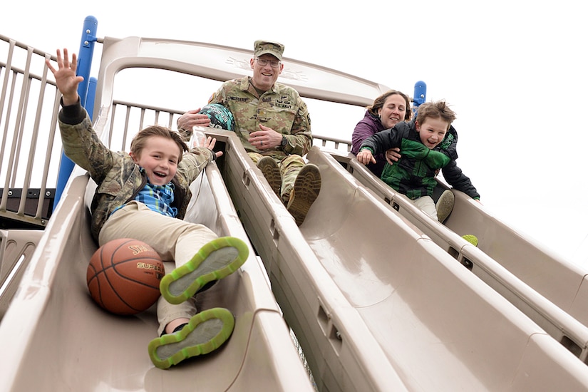 Maj. Trevor Wiegers, Mission Command Training Program observer coach/trainer and his wife Erin Wiegers, Fort Leavenworth Army Military Pay Office military pay technician, play after school with their children, Tristan (far left) and Trajan, at the Eisenhower Elementary School playground at Fort Leavenworth, Kansas March 22, 2024. A recent U.S. Army Financial Management Command initiative ensures AMPO employees who relocate with their service member spouses during permanent changes of station will maintain their positions within the command. (U.S. Army photo by Prudence Siebert)