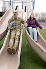 Maj. Trevor Wiegers, Mission Command Training Program observer coach/trainer, and Erin Wiegers, Fort Leavenworth Army Military Pay Office military pay technician, go down side-by-side slides together while playing with their children after school at the Eisenhower Elementary School playground at Fort Leavenworth, Kansas March 22, 2024. A recent U.S. Army Financial Management Command initiative ensures AMPO employees who relocate with their service member spouses during permanent changes of station will maintain their positions within the command. (U.S. Army photo by Prudence Siebert)