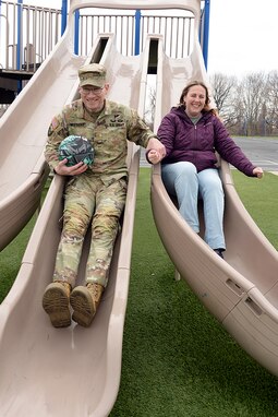 Maj. Trevor Wiegers, Mission Command Training Program observer coach/trainer, and Erin Wiegers, Fort Leavenworth Army Military Pay Office military pay technician, go down side-by-side slides together while playing with their children after school at the Eisenhower Elementary School playground at Fort Leavenworth, Kansas March 22, 2024. A recent U.S. Army Financial Management Command initiative ensures AMPO employees who relocate with their service member spouses during permanent changes of station will maintain their positions within the command. (U.S. Army photo by Prudence Siebert)