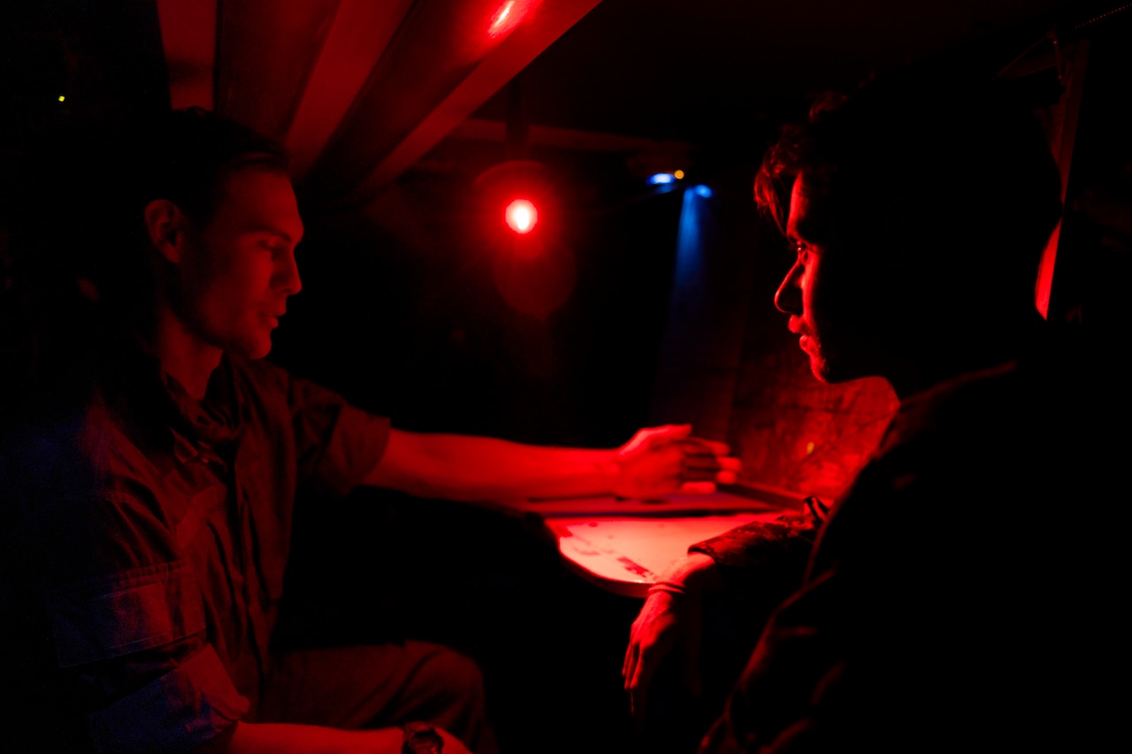 A service member in uniform sits at a table, illuminated by red light.