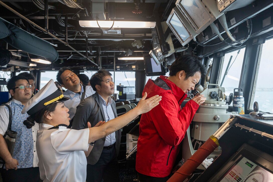 A Navy officer and a group of civilians gather on the bridge of a ship. One civilian looks through an optical device.
