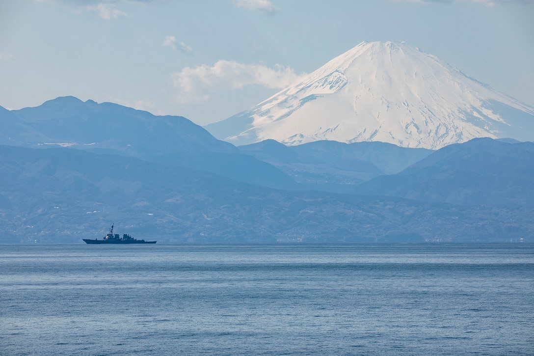 A military vessel sails with a mountain range in the background. The largest mountain is snow-capped.