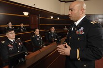 Five Army officers dressed in dark service uniforms are sitting in a dark wood -paneled room, in a jury box, and they are listening to comments being made by another bald Army officer dressed similarly who is facing them with his hands in front of him.