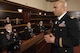 Five Army officers dressed in dark service uniforms are sitting in a dark wood -paneled room, in a jury box, and they are listening to comments being made by another bald Army officer dressed similarly who is facing them with his hands in front of him.