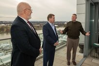 A man dressed in a dark green Army uniform jacket with lighter khaki pants is gesturing with this arms open as he talks towards two men dressed in suits. The are all standing on a rooftop terrace that overlooks a body of water.