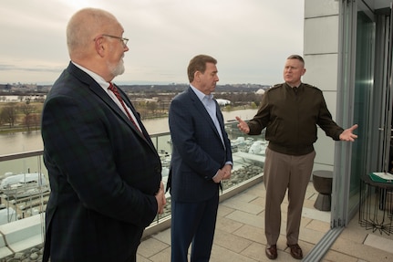 A man dressed in a dark green Army uniform jacket with lighter khaki pants is gesturing with this arms open as he talks towards two men dressed in suits. The are all standing on a rooftop terrace that overlooks a body of water.