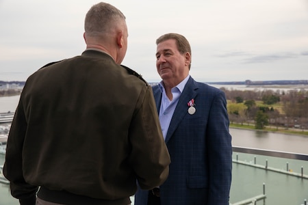 A man dressed in a dark suit with open collar is smiling and looking at another man dressed in a dark green Army uniform jacket, The man in the suit as a medal pinned to his lapel.