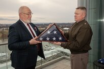 A man dressed in a dark suit with a white beard is holding a US flag that is folded into a wedge shape with only the dark blue background and white stars seen. Another man is holding the other corner of the same folded flag, and he is wearing a dark green Army uniform jack with lighter khaki pants. They are standing on the top floor terrace on top of a building.