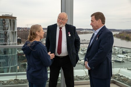 Two men dressed in dark suits are looking at a woman who is dressed in a dark hoodie and talking to them. They are standing on a terrace area on top of building that overlooks a body of water that has boats and another glass covered building in the background.