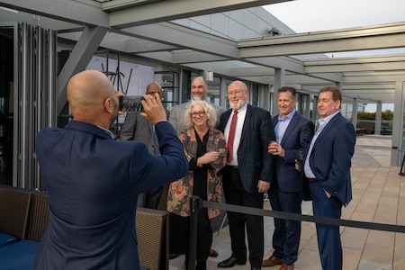 Several people dressed in coats and ties and dresses are posing for a photo as another person is holding up a cell phone to take the picture. They are outdoors on a patio with large grey support beams above them,