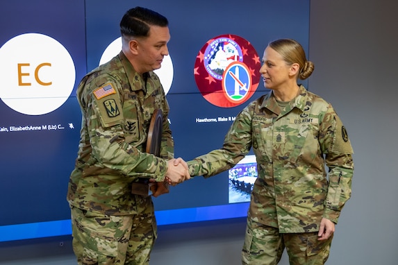 A man dressed in Army green camouflage uniform is sharking hands with a woman dressed similarly. They are standing in front of large screen monitors.