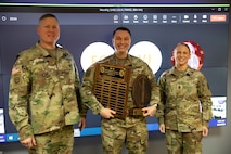 Two men and one woman dressed in Army green camouflage uniforms are posing for a picture while standing in front of a large screen monitor. The man in the middle has wooden award plaques in his hands.