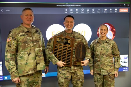 Two men and one woman dressed in Army green camouflage uniforms are posing for a picture while standing in front of a large screen monitor. The man in the middle has wooden award plaques in his hands.