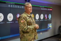 A man dressed in Army green camouflage uniform is speaking in front of large screen monitors. He has his hands together in front of his body.