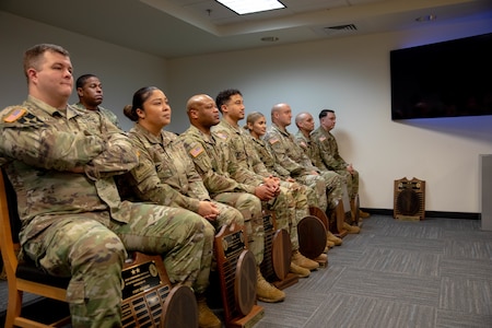 About ten Army soldiers dressed in green camouflage unifroms are seated and are looking towards the right side of the picture. In front of several on the floor are large wooden award plaques at their feet.