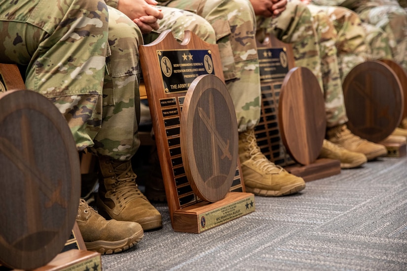 Several large wooden award plaques resting on the floor by the legs of Army Soldiers dressed in green camouflage uniforms. Only their lower legs and boots are visible in this picture.