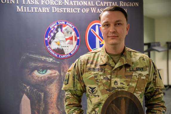 A man dressed in Army green camouflage uniforms is posing with a large wooden award plaque in his hands. Behind him is a poster that reads Joint Task Force-National Capital Region Military District of Washington.