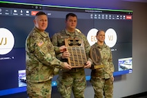 A man dressed in Army green camouflage uniforms is handing another man dressed similarly and standing to his left a large wooden award plaque. A woman dressed in Army green camouflage uniforms is handing the man another plaque and she is standing on his left.