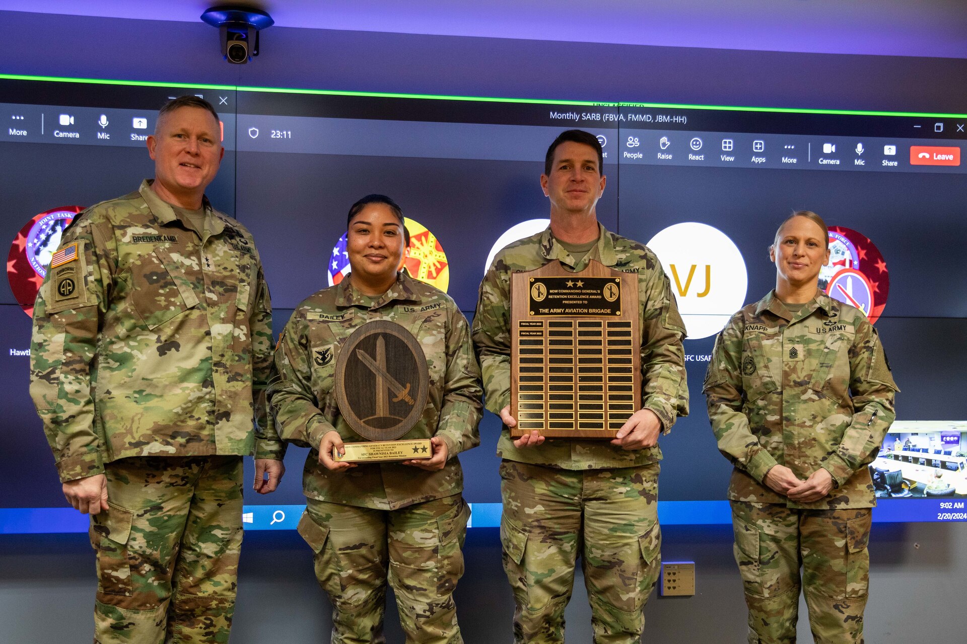 Two men and two women dressed in Army green camouflage uniforms are posing for a picture while standing in front of a large screen monitor. The two middle people have wooden award plaques in their hands.