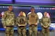 Two men and two women dressed in Army green camouflage uniforms are posing for a picture while standing in front of a large screen monitor. The two middle people have wooden award plaques in their hands.