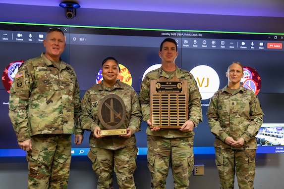 Two men and two women dressed in Army green camouflage uniforms are posing for a picture while standing in front of a large screen monitor. The two middle people have wooden award plaques in their hands.