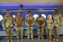 Six people dressed in Army green camouflage uniforms are posing for a picture while standing in front of a large screen monitor. The three middle people have wooden award plaques in their hands.