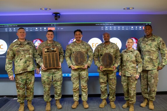 Six people dressed in Army green camouflage uniforms are posing for a picture while standing in front of a large screen monitor. The three middle people have wooden award plaques in their hands.