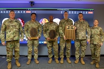 Several people dressed in Army green camouflage uniforms are posing for a picture while standing in front of a large screen monitor. Some people have wooden award plaques in their hands.