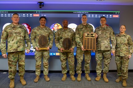 Several people dressed in Army green camouflage uniforms are posing for a picture while standing in front of a large screen monitor. Some people have wooden award plaques in their hands.