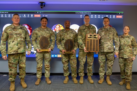 Several people dressed in Army green camouflage uniforms are posing for a picture while standing in front of a large screen monitor. Some people have wooden award plaques in their hands.
