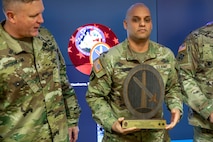 A man dressed in Army green camouflage uniforms is posing for a picture while standing in front of a large screen monitor. He has a wooden award plaque in his hands. Another man in Army green camouflage is looking at the award and speaking.