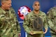 A man dressed in Army green camouflage uniforms is posing for a picture while standing in front of a large screen monitor. He has a wooden award plaque in his hands. Another man in Army green camouflage is looking at the award and speaking.