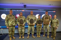 Six people dressed in Army green camouflage uniforms are posing for a picture while standing in front of a large screen monitor. Some people have wooden award plaques in their hands.