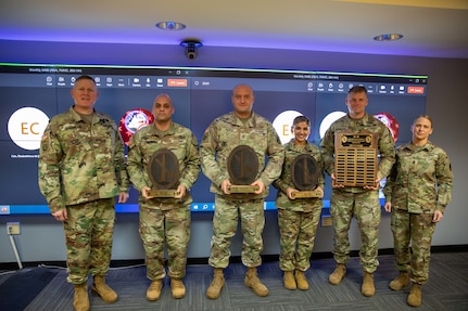 Six people dressed in Army green camouflage uniforms are posing for a picture while standing in front of a large screen monitor. Some people have wooden award plaques in their hands.
